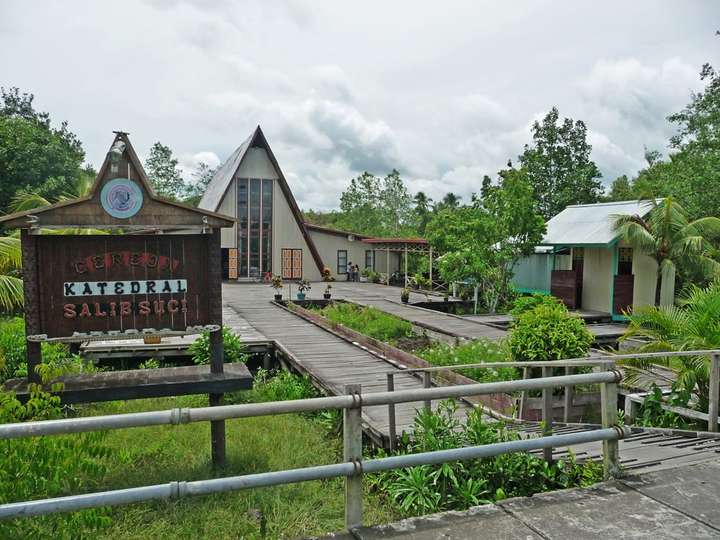 Papan nama berukir "Katedral Salib Suci" di persimpangan titian kayu, di antara pepohonan hijau