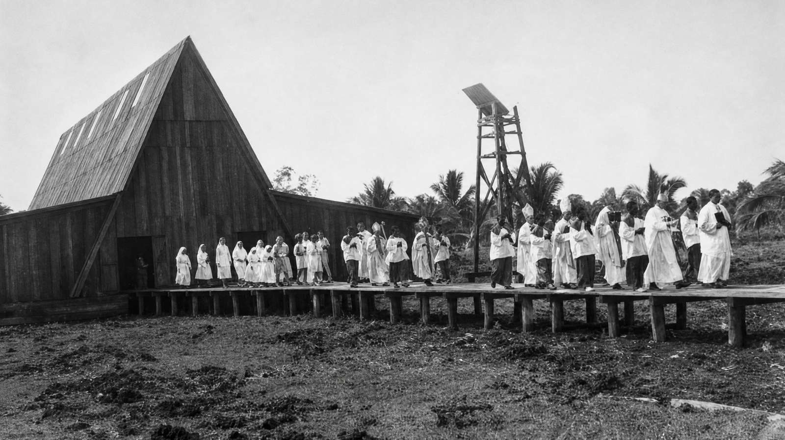 Black-and-white photograph: a procession of clergy in white vestments on the boardwalk before the A-frame church and bell tower