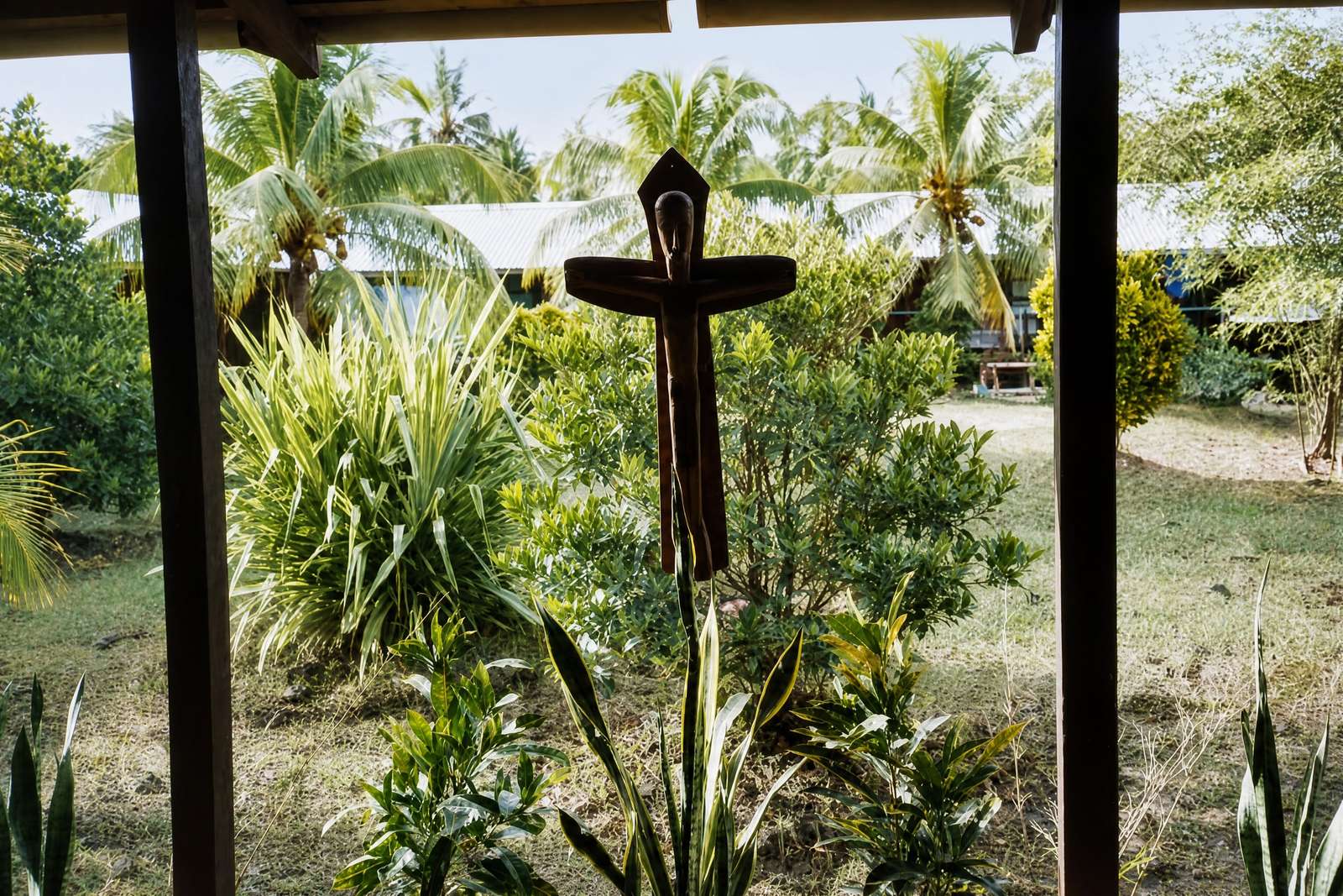 A dark wooden cross in a tropical garden, seen through a window frame